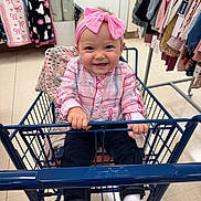 Lola is registered to the contest to win money with this photo: baby, child, shopping_cart, pink_bow, headband, plaid_shirt, dark_pants, white_socks, indoor, store, clothing_rack, blanket, happy, smiling, holding, sitting, retail, cute, floor, person