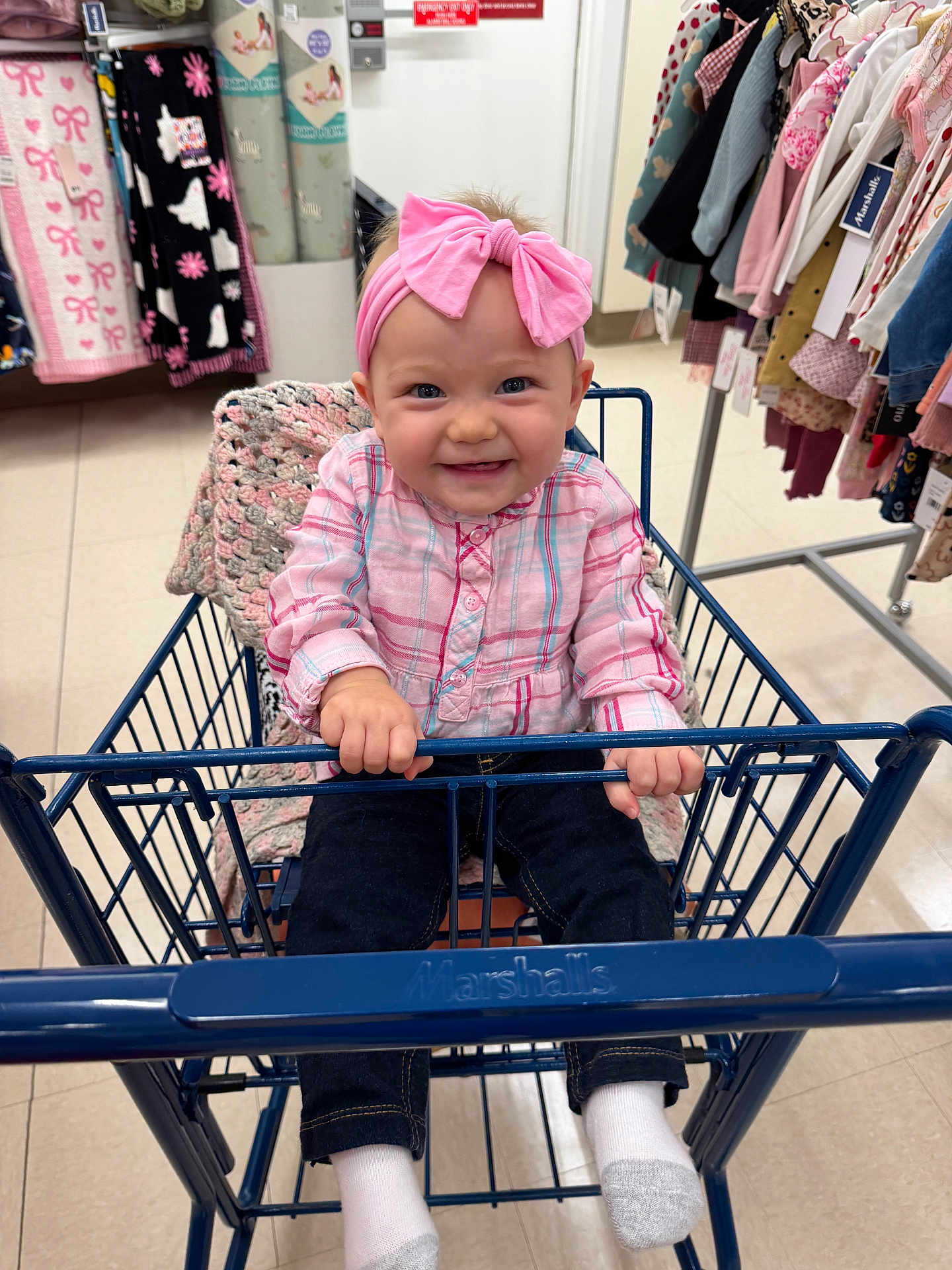 Lola is registered to the contest to win money with this photo: baby, child, shopping_cart, pink_bow, headband, plaid_shirt, dark_pants, white_socks, indoor, store, clothing_rack, blanket, happy, smiling, holding, sitting, retail, cute, floor, person