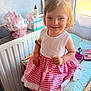 baby_bottle, chair, child, cushion, dress, girl, hair, hair_tie, happy, indoors, makeup_palette, person, personal_items, portrait, radiator, red_checkered, smiling, sunlight, white_top, window