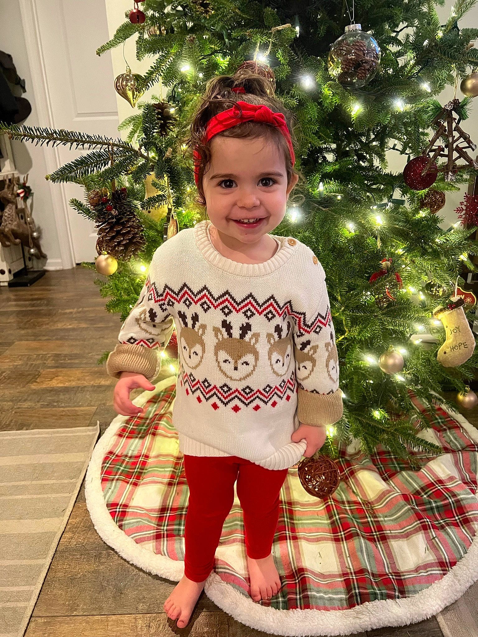 Eliana is registered to the contest to win money with this photo: baby_toddler_clothing, christmas, christmas_decoration, christmas_ornament, christmas_tree, event, happy, headwear, holiday, holiday_ornament, human_body, joy, ornament, pattern, person, plaid, red, sleeve, smile, standing