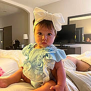 Carina joined the competition — help win amazing prizes! baby, child, infant, blue_dress, headband, bow, bed, adult_arm, indoor, curious_look, wide_eyes, soft_lighting, mirror, television, furniture, skin, legs, hand, person, room