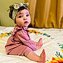 child, baby, infant, toddler, headband, curly_hair, big_eyes, sitting, bed, blanket, floral_pattern, pink_clothing, portrait, indoors, soft_lighting, cute_expression, toes, feet, fashion_headwrap, ruffled_outfit