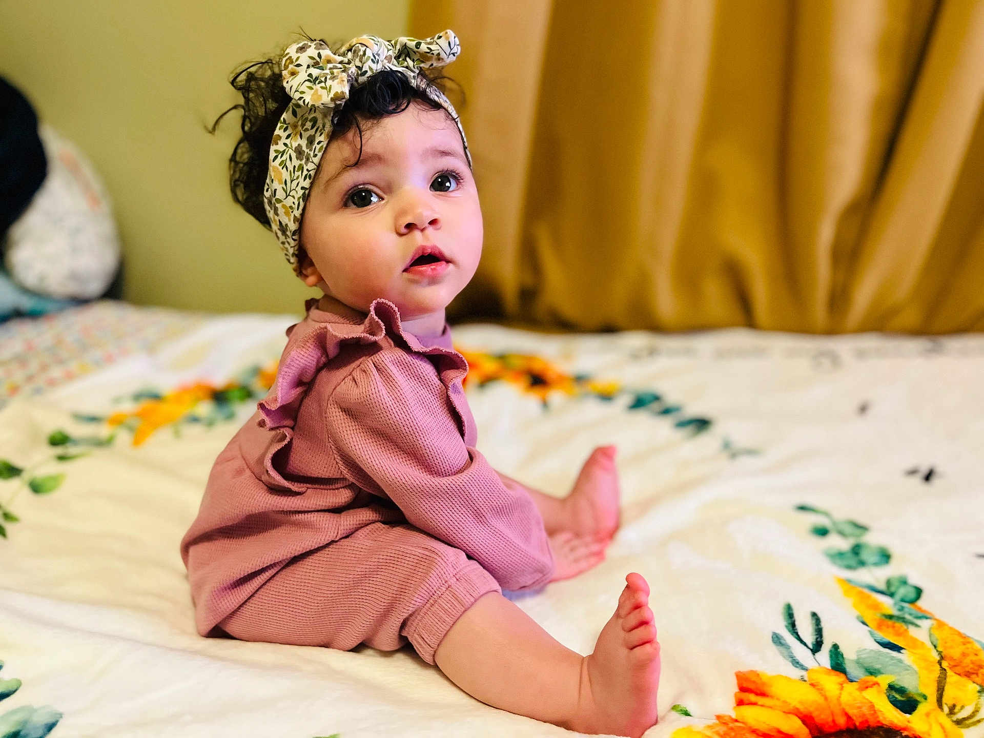 Izabella is registered to the contest to win money with this photo: child, baby, infant, toddler, headband, curly_hair, big_eyes, sitting, bed, blanket, floral_pattern, pink_clothing, portrait, indoors, soft_lighting, cute_expression, toes, feet, fashion_headwrap, ruffled_outfit
