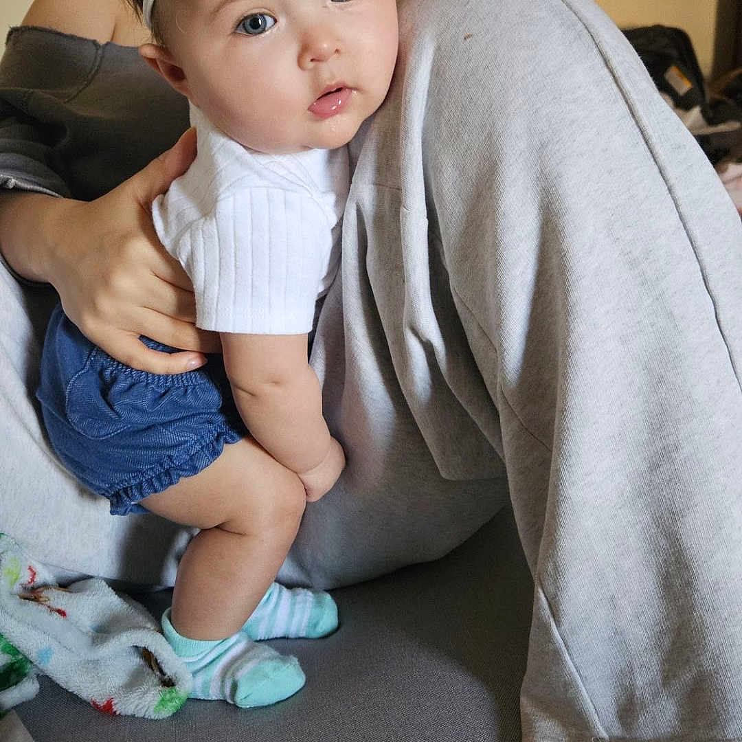 Leila is registered to the contest to win money with this photo: baby, blanket, blue_eyes, blue_shorts, child, couch, cute, expression, face, flower, gray_pants, hand, headband, holding, home, indoor, person, sitting, socks, white_shirt