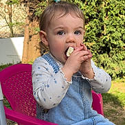 Giulia a rejoint le concours — aidez-le/la à gagner de superbes lots ! child, toddler, girl, eating, snack, hands, face, eyes, hair_clip, plastic_chair, denim_dress, overalls, outdoor, garden, trees, portrait, sitting, pink, cute, food