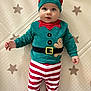 baby, blue_eyes, child, cute, elf_costume, festive_clothing, gingerbread_man, green_shirt, holiday, indoors, infant, lying_down, mat, person, portrait, red_hat, smiling, soft_lighting, star_pattern, striped_pants