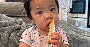 Kai Yumi is registered to the contest to win money with this photo: toddler, child, baby, indoor, kitchen, freeze_pop, blanket, sofa, curly_hair, pink_onesie, person, cute, eating, snack, modern_kitchen, appliance, cabinet, stainless_steel, home, cozy