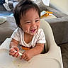 baby, child, toddler, smiling, laughing, face, portrait, high_chair, feeding, food, spoon, white_shirt, dark_hair, indoor, living_room, sofa, cushion, tray, messy_eating, happy
