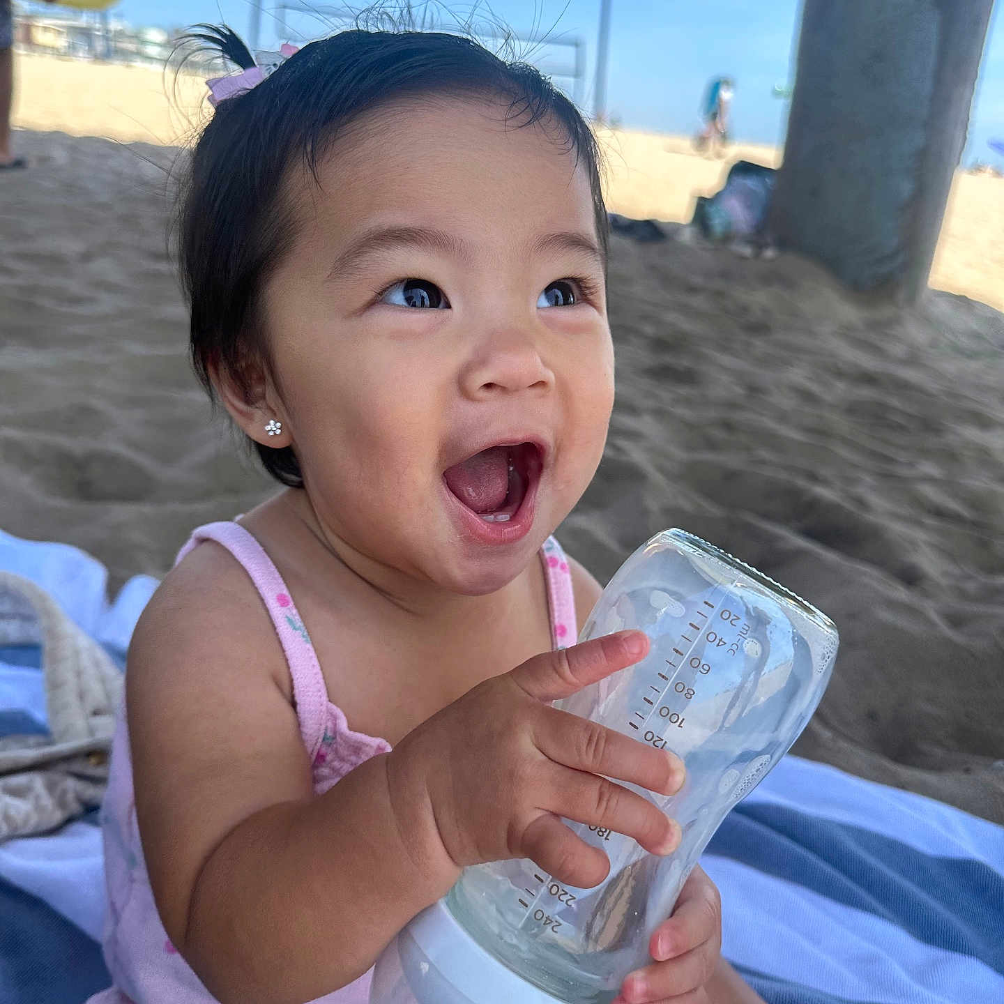 Kai Yumi is registered to the contest to win money with this photo: baby, child, girl, bottle, beach, sand, smile, happy, towel, outdoor, sunny, earrings, hair_tie, pink_clothing, person, sitting, daytime, portrait, cute, joyful