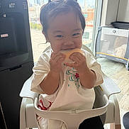 Kai Yumi is registered to the contest to win money with this photo: black_shorts, child, daylight, eating, food, happy, high_chair, home, indoors, kitchen, pigtails, playful, portrait, sitting, smiling, snack, sunlight, toddler, white_shirt, window
