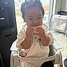 child, toddler, smiling, eating, snack, food, high_chair, pigtails, indoors, window, sunlight, portrait, sitting, white_shirt, black_shorts, home, kitchen, daylight, happy, playful