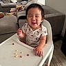 toddler, child, high_chair, food, smile, dimples, hello_kitty, shirt, indoor, living_room, sofa, toy, couch, floor, wood_floor, happy, person, furniture, cute, eating