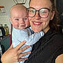 Benjamin is registered to the contest to win money with this photo: baby, woman, glasses, smile, indoor, kitchen, holding, person, face, hand, clothing, appliance, shelf, plate, countertop, bottle, light, portrait, happy, home