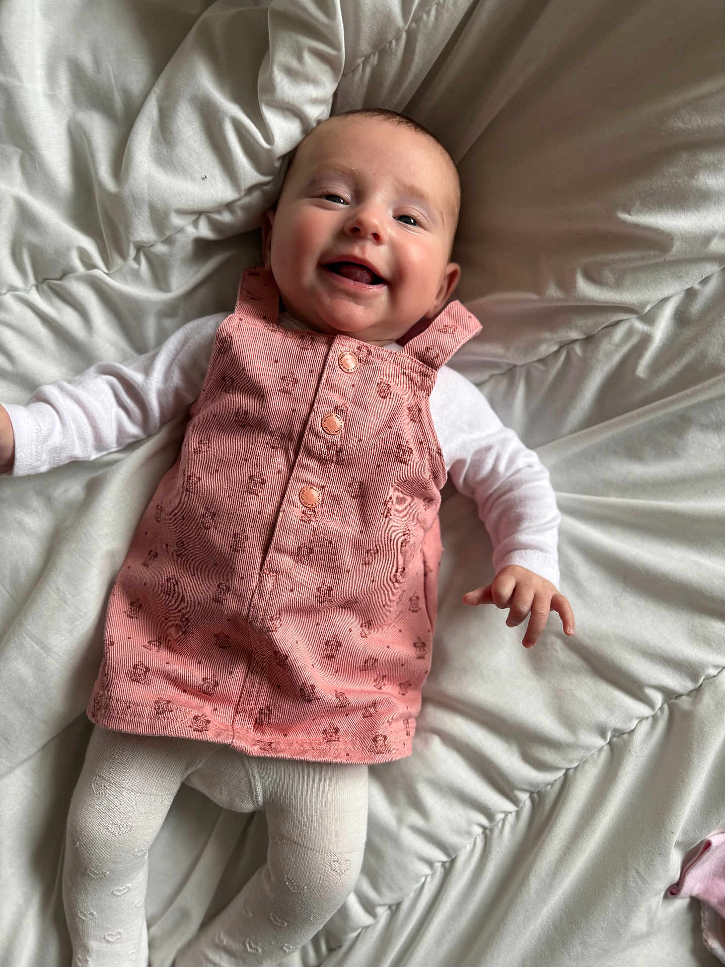 Lou a rejoint le concours — aidez-le/la à gagner de superbes lots ! baby, smiling, pink_dress, white_tights, lying_down, bed, comforter, infant, happy, child, portrait, cute, indoors, soft_lighting, fabric, clothing, person, face, hand, head