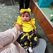 Autumn is registered to the contest to win money with this photo: baby, child, yellow_clothing, headband, sunflower_pants, socks, person, indoor, carpet, cabinet, gift_bag, seated, lap, curious, expression, floor, furniture, holding_hand, casual, portrait