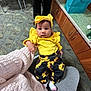 baby, child, yellow_clothing, headband, sunflower_pants, socks, person, indoor, carpet, cabinet, gift_bag, seated, lap, curious, expression, floor, furniture, holding_hand, casual, portrait