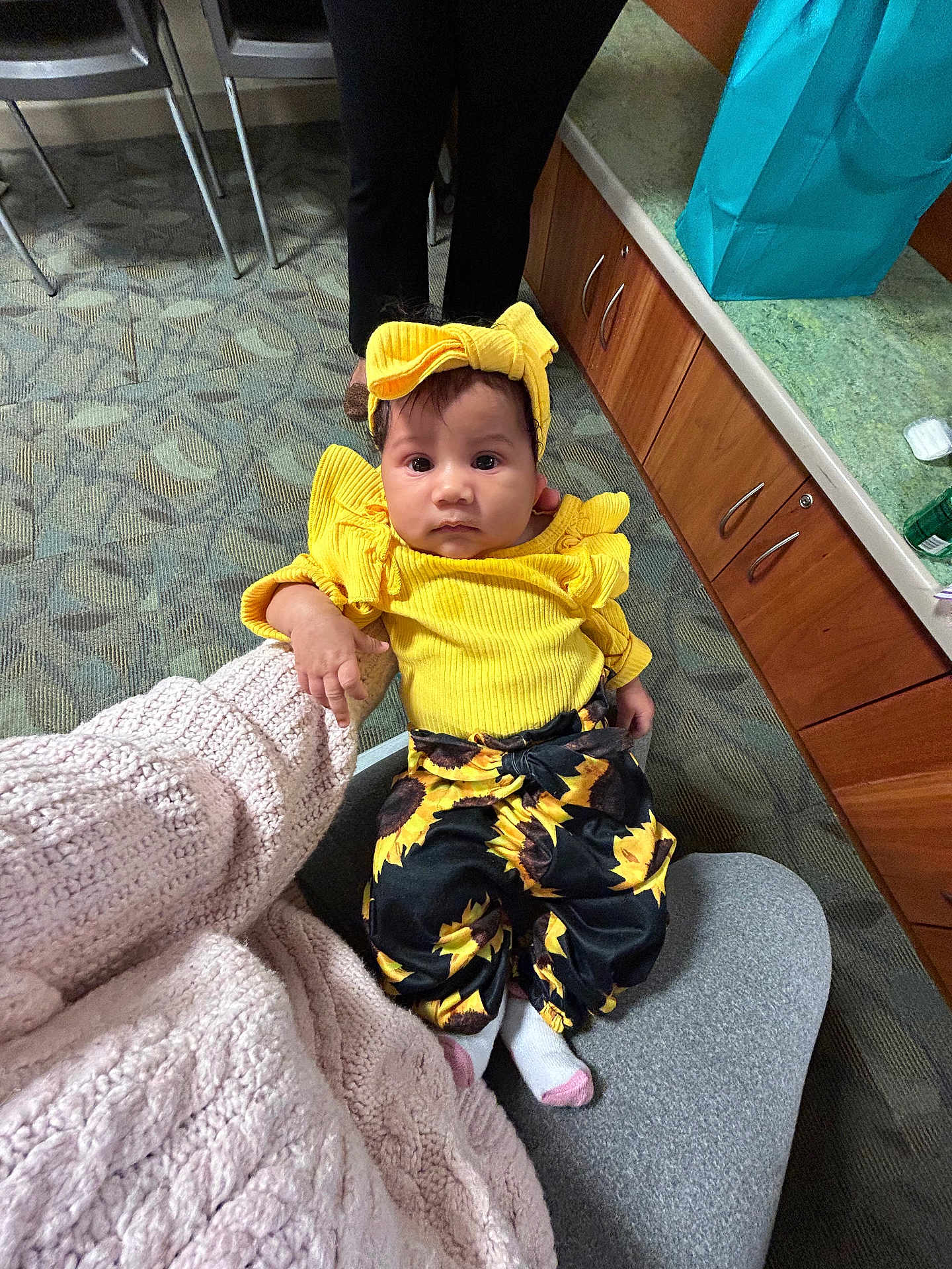 Autumn is registered to the contest to win money with this photo: baby, child, yellow_clothing, headband, sunflower_pants, socks, person, indoor, carpet, cabinet, gift_bag, seated, lap, curious, expression, floor, furniture, holding_hand, casual, portrait