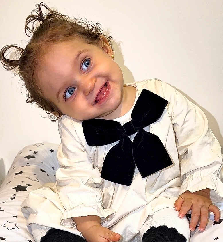 Hailey participe au concours pour gagner de l'argent avec cette photo : toddler, child, blue_eyes, smile, pigtails, black_bow, cream_dress, sitting, star_pattern, cushion, indoor, cute, happy, portrait, young_child, baby, playful, head_tilt, small_ears, fingertips