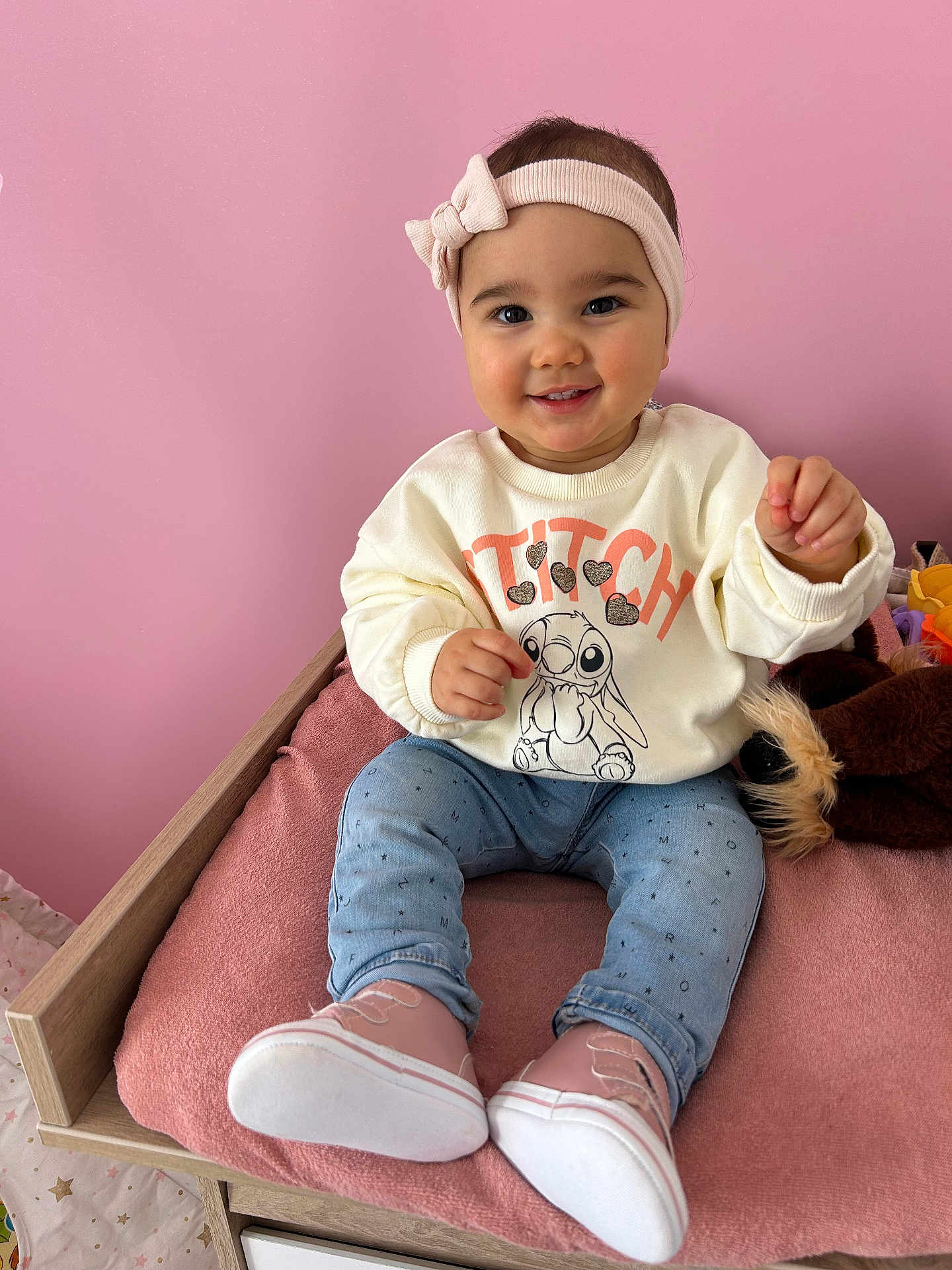 Lyana participe au concours pour gagner de l'argent avec cette photo : child, toddler, baby, headband, pink_background, smile, sitting, shoes, sweatshirt, jeans, cushion, plush_toy, wooden_bed, nursery, cozy, portrait, rosy_cheeks, indoors, cute, happy