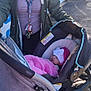 woman, baby, stroller, car_seat, blanket, outdoor, pavement, keys, necklace, coat, hat, pink_blanket, infant, smile, parent, lanyard, sunlight, fence, sidewalk, urban_scene