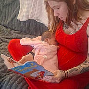 Serenity is registered to the contest to win money with this photo: mother, infant, baby, breastfeeding, book, reading, red_dress, tattoo, sofa, cushion, blanket, cozy, living_room, carpet, hand, portrait, face, woman, child, parent