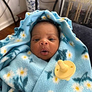 Kennedi is registered to the contest to win money with this photo: baby, blanket, blue, flower_pattern, pacifier, indoor, face, infant, curious_expression, soft_texture, cozy, furniture, poster, wood_floor, table, decor, person, portrait, wrapped, child
