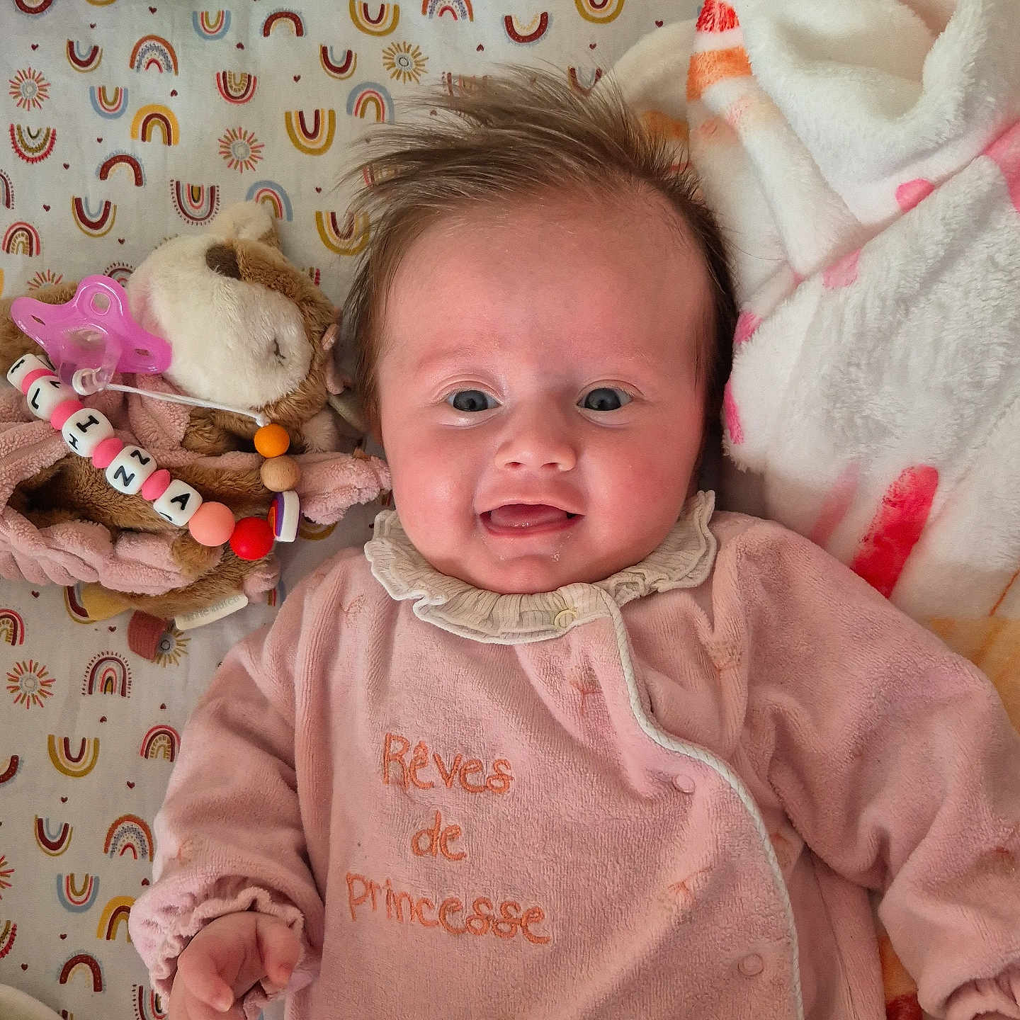 Mélina participe au concours pour gagner de l'argent avec cette photo : baby, beaded_clip, blanket, cute, drool, embroidered_text, hand, infant, lying_down, nursery, pacifier, pink_onesie, plush_toy, portrait, rainbow_pattern, rosy_cheeks, sheet, smiling, soft_toy, tousled_hair