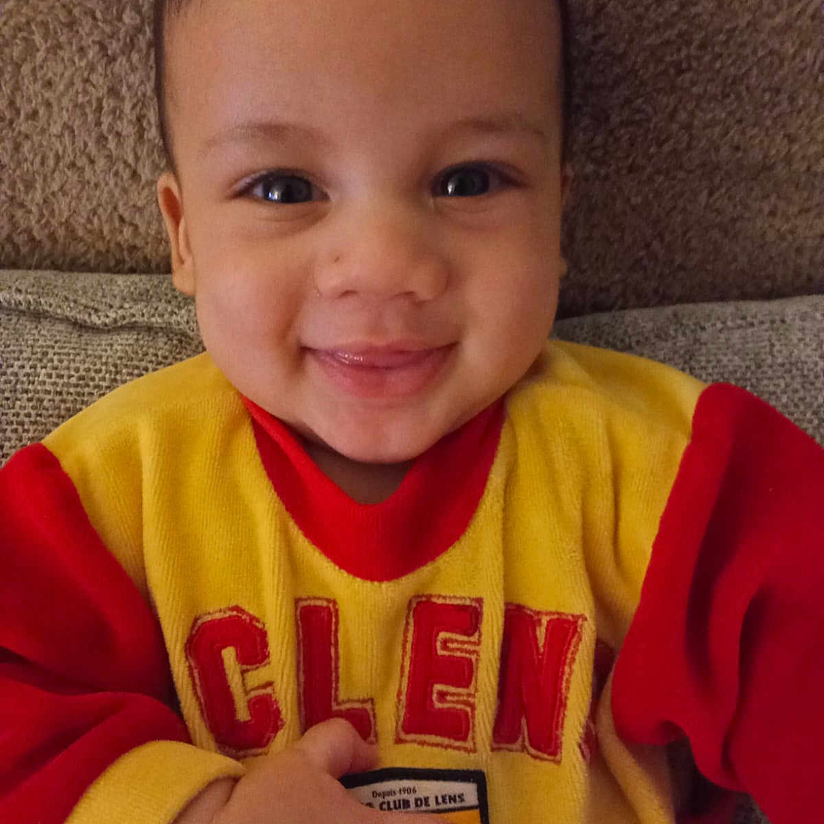 Prince Joseph a rejoint le concours — aidez-le/la à gagner de superbes lots ! baby, casual, child, closeup, comfort, couch, cute, face, hand, happy, indoors, infant, person, portrait, red_clothing, seated, smile, sweater, warm_colors, yellow_clothing