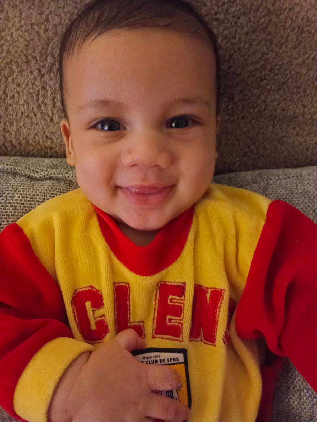 Prince Joseph a rejoint le concours — aidez-le/la à gagner de superbes lots ! baby, child, smile, face, sweater, yellow_clothing, red_clothing, couch, seated, happy, portrait, indoors, person, infant, hand, comfort, cute, warm_colors, closeup, casual