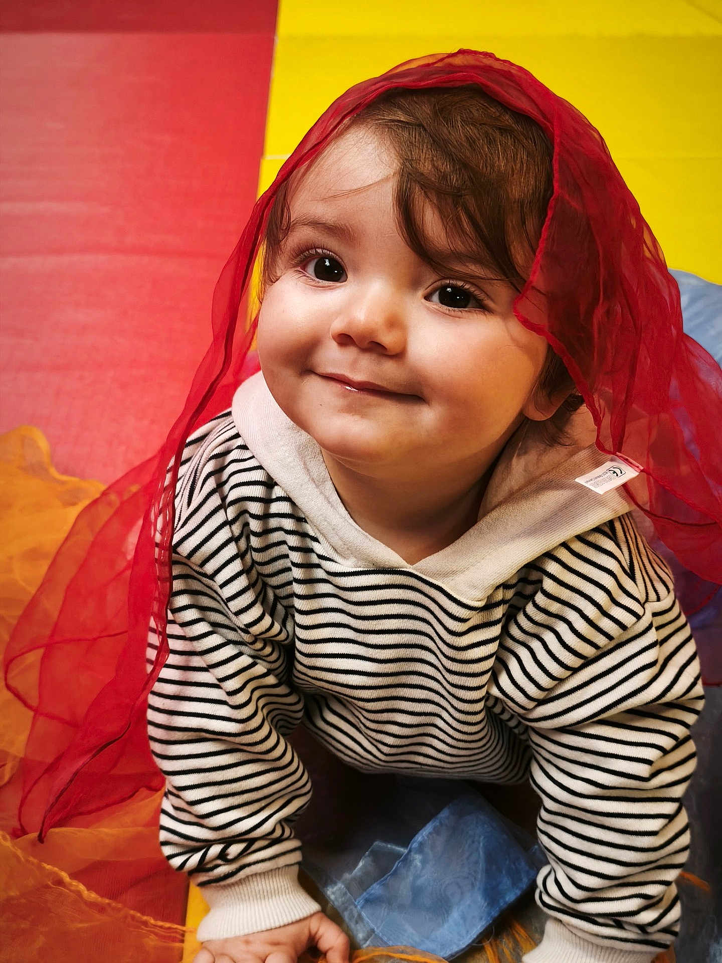 Nahel participe au concours pour gagner de l'argent avec cette photo : toddler, child, smile, face, striped_clothing, hoodie, red_scarf, crawling, colorful_background, yellow, red, blue, playmat, curly_hair, cute, indoors, happy, portrait, baby, person