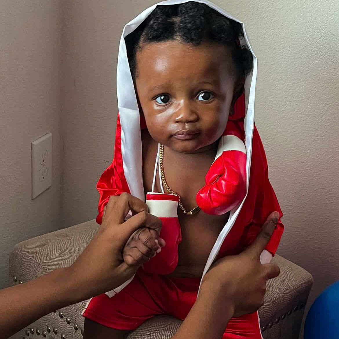 Zyaire joined the competition — help win amazing prizes! baby, child, portrait, sitting, red_outfit, hood, gold_chain, fashion, indoors, living_room, chair, adults, hands, dark_skin, curly_hair, serious_expression, photography, home, outfit, full_body