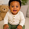 baby, child, smiling, curly_hair, sitting, white_socks, green_pants, cream_top, teddy_bear, plush_toy, carpet, indoor, portrait, happy, cute, young_child, person, home, cozy, soft_lighting