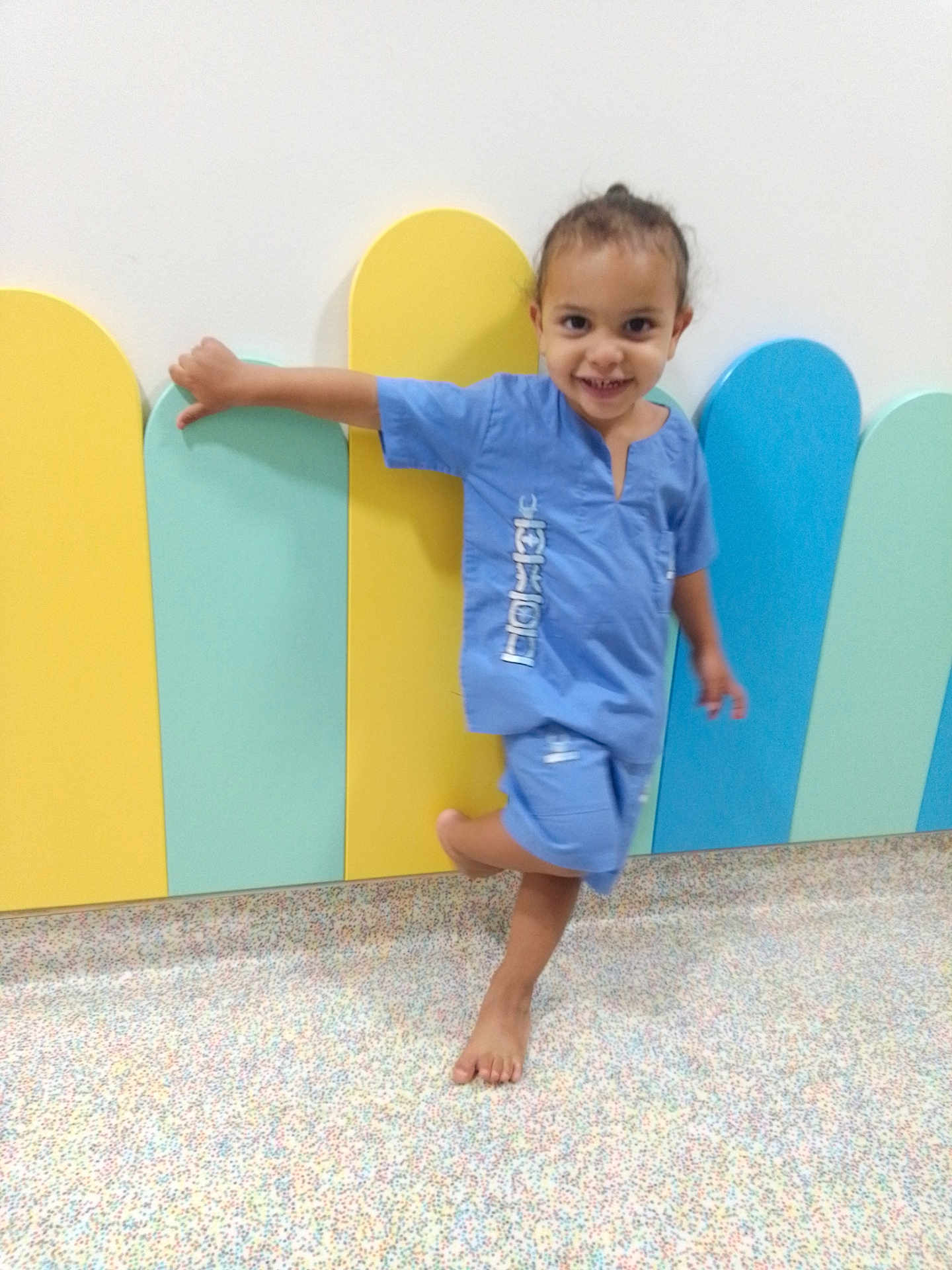 Jilyan a rejoint le concours — aidez-le/la à gagner de superbes lots ! child, smiling, barefoot, blue_clothing, scrubs, colorful_background, pastel_colors, playful, happy, indoor, wall, floor, person, cute, young, posing, joyful, small_child, casual, fun
