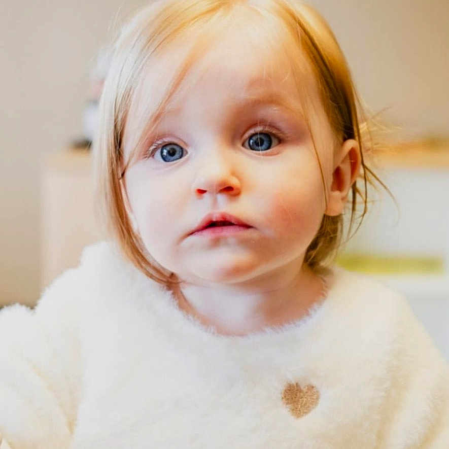 Alba participe au concours pour gagner de l'argent avec cette photo : baby, blonde_hair, blue_eyes, child, close_up, curious, cute, expression, face, heart_detail, indoor, innocent, person, portrait, soft_lighting, sweater, toddler, white_clothing, young, young_child