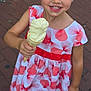 child, confection, daylight, dress, face, food, footwear, girl, hair_clips, hand, happy, ice_cream, outdoor, patterned_dress, pavement, person, sandals, smile, summer, sweet