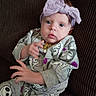 baby, bow, cartoon, child, clothing, couch, cute, expression, eyes, face, furniture, hand, headwear, indoor, infant, onesie, pattern, portrait, seated, young