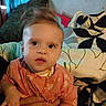 Myla is registered to the contest to win money with this photo: adult_hand, baby, bed, bedding, bedroom, blanket, child, curtain, eyes, face, indoor, infant, necklace, orange_clothing, patterned_fabric, pillow, portrait, sitting, support, wide_eyed
