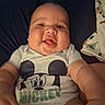 baby, child, mickey_mouse, onesie, smiling, tongue_out, chubby_cheeks, infant, cute, lying_down, blanket, animal_print, blue_background, closeup, face, happy, person, indoors, soft_lighting, casual_clothing