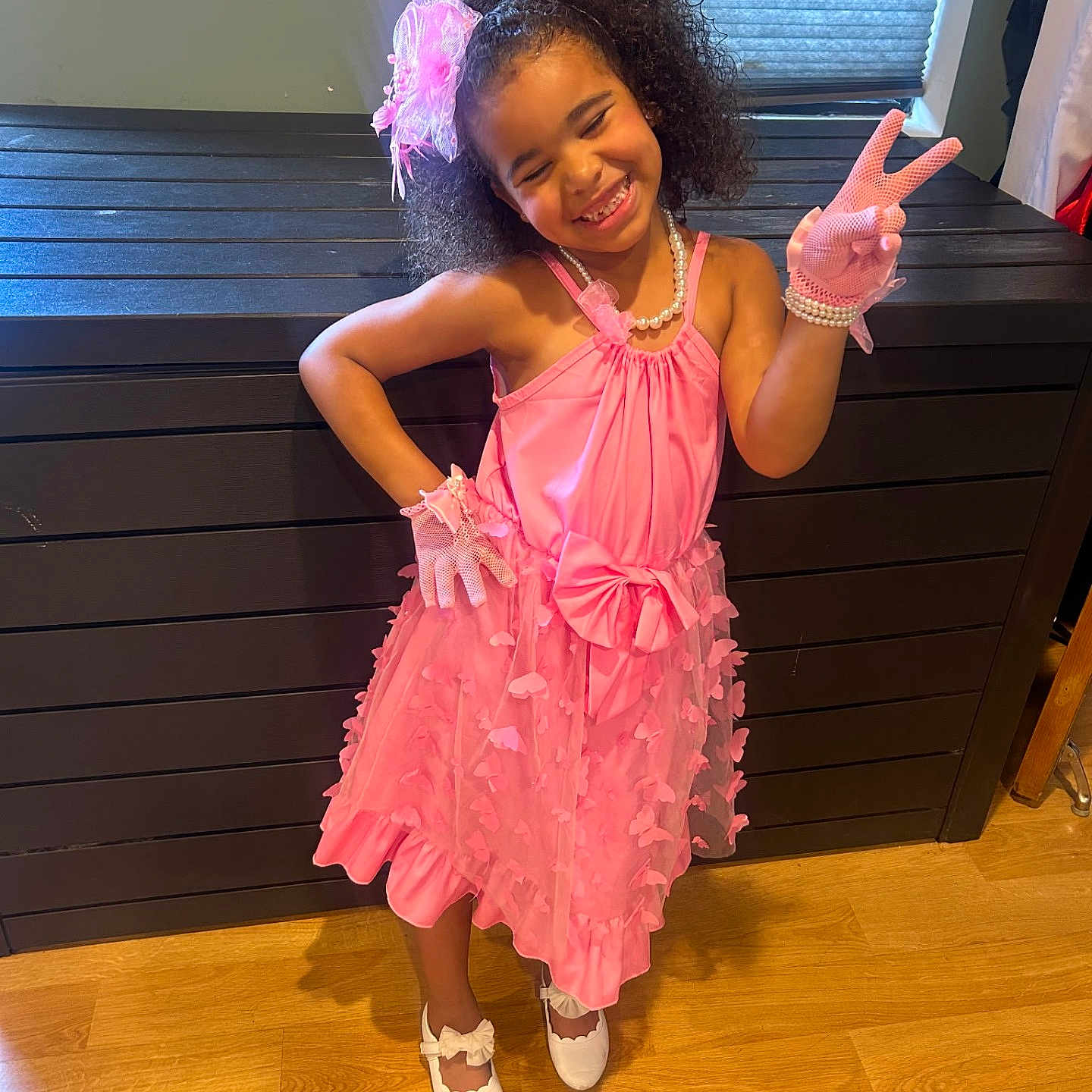 Nevaeh is registered to the contest to win money with this photo: child, girl, pink_dress, gloves, smile, peace_sign, curly_hair, headband, pearl_necklace, white_shoes, wooden_floor, cabinet, window, indoor, fashion, happy, posing, accessories, playful, confident