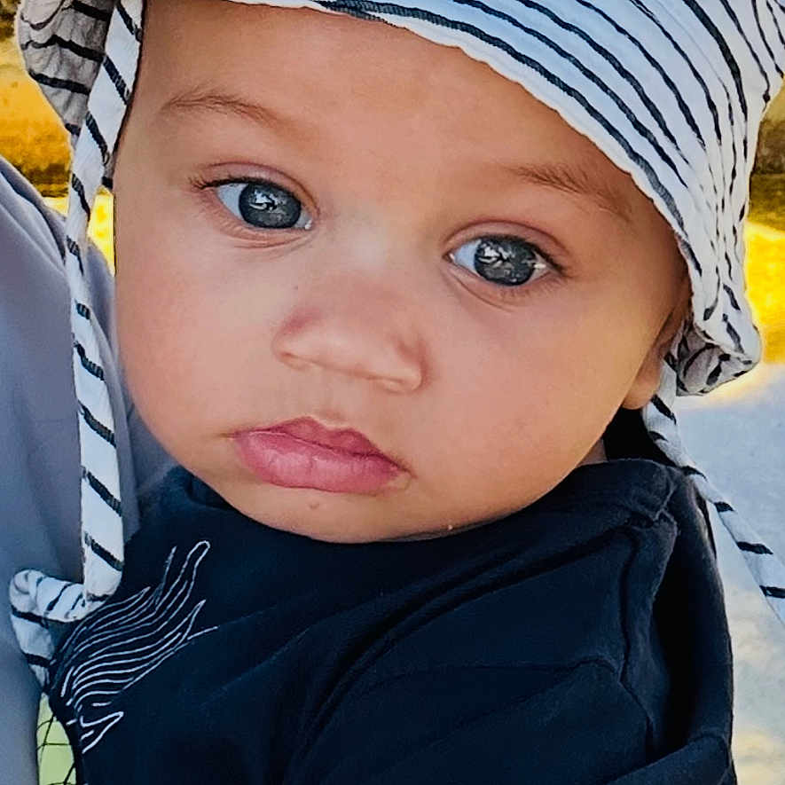 Ellis a rejoint le concours — aidez-le/la à gagner de superbes lots ! baby, black_shirt, child, close_up, cute, daylight, expression, eyes, face, grass, hat, headwear, outdoor, person, portrait, skin, striped_hat, summer, toddler, tree