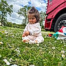casual_clothing, child, cloudy_sky, curious, cute, daisy, daytime, field, flower, grass, greenery, nature, outdoor, playful, red, sitting, toddler, tree, truck, vehicle