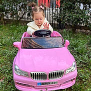 Aria a rejoint le concours — aidez-le/la à gagner de superbes lots ! child, toddler, toy_car, pink_car, outdoor, grass, greenery, fence, pigtails, bows, cute, playing, happy, person, young_child, steering_wheel, sweater, nature, daylight, portrait