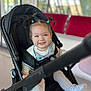 toddler, child, smiling, stroller, black_stroller, white_shoes, denim_overall, pigtails, happy, indoor, seated, cute, person, baby, clothing, footwear, portrait, infant, young_child, casual