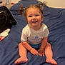 toddler, child, smiling, sitting, bed, bedspread, indoor, pigtails, hair_ties, clothing, white_tshirt, blue_shorts, happy, face, person, room, bag, spray_bottle, casual, home
