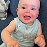 Bryer is registered to the contest to win money with this photo: baby, blue, child, close_up, clothing, couch, eyes, face, hands, happy, indoor, infant, living_room, patterned_pillow, portrait, romper, sitting, smiling, sofa, toy