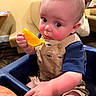 adult, baby, background, bib, blue_clothing, child, citrus, eating, food, fruit, high_chair, indoor, infant, orange, overalls, portrait, restaurant, seated, table, wide_eyed