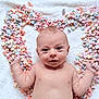 baby, infant, newborn, conversation_hearts, candy, heart_shape, blanket, pastel_colors, lying_down, portrait, bare_chest, cute, eyes, hands, smile, soft_texture, valentine, close_up, playful, props