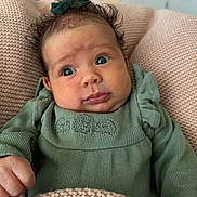 Nora joined the competition — help win amazing prizes! baby, infant, child, face, portrait, green_clothing, bow, blanket, knitted_blanket, hand, cute, expression, indoors, cozy, soft_texture, curly_hair, small_person, sitting, young, person