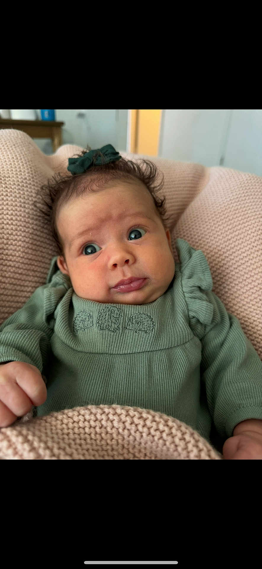 Nora joined the competition — help win amazing prizes! baby, infant, child, face, portrait, green_clothing, bow, blanket, knitted_blanket, hand, cute, expression, indoors, cozy, soft_texture, curly_hair, small_person, sitting, young, person