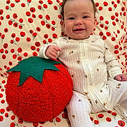 Nora is registered to the contest to win money with this photo: baby, child, onesie, plush_toy, tomato_pillow, red, green, white, pattern, cherry_pattern, blanket, smile, happy, sitting, indoor, cute, face, hand, hair, soft_toy